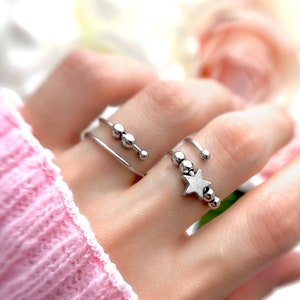 May include: A silver ring with a star charm and a row of small beads. The ring is on a finger wearing a simple silver band.