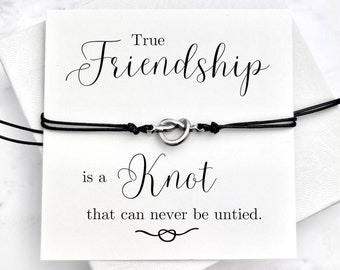 Puede incluir: Una tarjeta blanca con texto negro que dice "True Friendship is a Knot that can never be untied." Una pulsera de cuerda negra con un dije de nudo plateado está encima de la tarjeta.