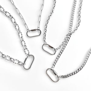 May include: Four silver-toned metal necklaces, each featuring a distinct chain link style and a central oval-shaped carabiner clasp. The chains vary in texture, including smooth elongated links, textured round links, and standard curb chain links.