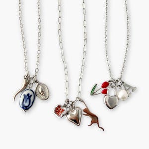 May include: Three silver necklaces with various charms. One necklace has a blue and white pendant, a horn, and a silver oval charm. Another has a heart, a cat, and a heart-shaped charm. The third has a heart-shaped lock, a pearl, and a cherry charm.