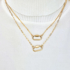 May include: A layered gold-tone necklace featuring two delicate chains of varying lengths. Each chain is accented with a small, polished oval carabiner-style clasp link centered as a focal point, resting against a white ribbed fabric texture.