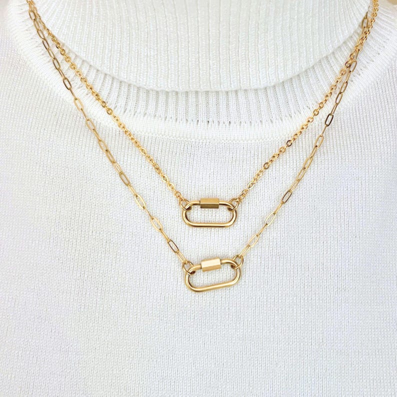 May include: A layered gold-tone necklace featuring two delicate chains of varying lengths. Each chain is accented with a small, polished oval carabiner-style clasp link centered as a focal point, resting against a white ribbed fabric texture.