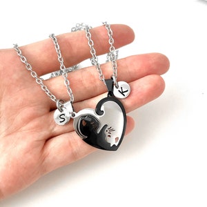 May include: Two silver necklaces with a black and white heart pendant. The pendant is split in half, with a black cat on one side and a white cat on the other. Each necklace has a small silver disc with a letter on it, one says 'S' and the other says 'K'.
