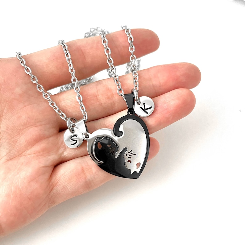 May include: Two silver necklaces with a black and white heart pendant. The pendant is split in half, with a black cat on one side and a white cat on the other. Each necklace has a small silver disc with a letter on it, one says 'S' and the other says 'K'.