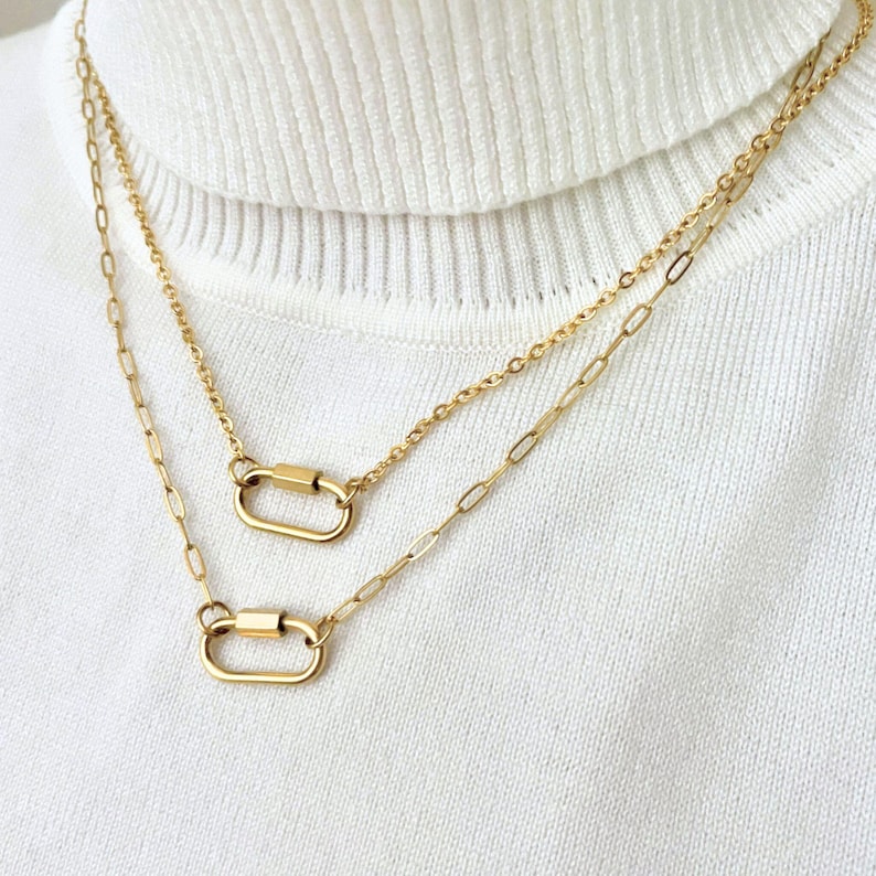 May include: A double-layered gold necklace featuring two delicate chains with elongated oval links. Each chain is connected to a small, polished gold carabiner-style clasp that serves as a central focal point on the front of the necklace.