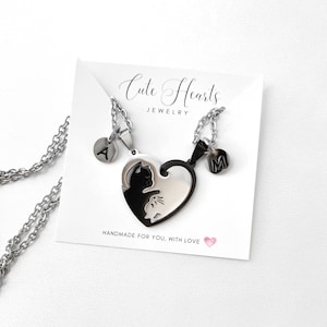 May include: Two silver necklaces with heart-shaped pendants. The pendants are black and white and feature a cat silhouette. Each necklace has a small silver charm with a letter on it. The charms say 'A' and 'M'. The necklaces are displayed on a white card with the text 'Cute Hearts Jewelry' and 'Handmade for you, with love'. 