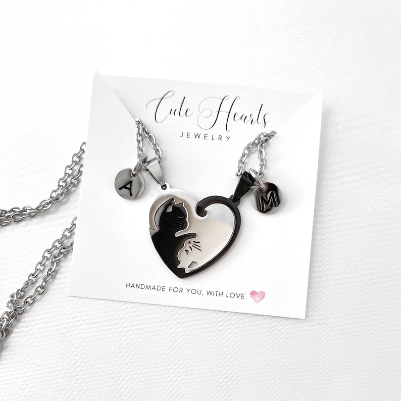 May include: Two silver necklaces with heart-shaped pendants. The pendants are black and white and feature a cat silhouette. Each necklace has a small silver charm with a letter on it. The charms say 'A' and 'M'. The necklaces are displayed on a white card with the text 'Cute Hearts Jewelry' and 'Handmade for you, with love'. 