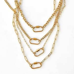 Puede incluir: Cuatro collares dorados con diferentes estilos de cadena. Cada collar presenta un cierre grande en forma ovalada. Las cadenas incluyen estilos de eslabones, clip y cuerda, creando un aspecto en capas. Las joyas se exhiben sobre un fondo blanco.