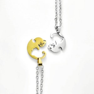 May include: Two gold and silver necklaces with matching pendants. The pendants are shaped like cats facing each other, with one cat's tail forming the other cat's head. The necklaces are on a white background.