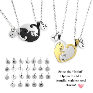 May include: Two silver necklaces with matching pendants. One pendant is a black and white yin yang symbol with two cats facing each other. The other pendant is a gold and silver heart with two cats facing each other. Both pendants have a small circle with a letter on it. The letters are J, M, A, and E. Below the necklaces is a chart of letters A through Z in silver circles. The text below the chart reads 'Select the 'Initial' Option to add 2 beautiful stainless steel charms!' with a pink heart icon.
