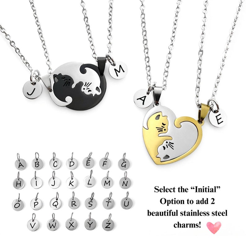 May include: Two silver necklaces with matching pendants. One pendant is a black and white yin yang symbol with two cats facing each other. The other pendant is a gold and silver heart with two cats facing each other. Both pendants have a small circle with a letter on it. The letters are J, M, A, and E. Below the necklaces is a chart of letters A through Z in silver circles. The text below the chart reads 'Select the 'Initial' Option to add 2 beautiful stainless steel charms!' with a pink heart icon.