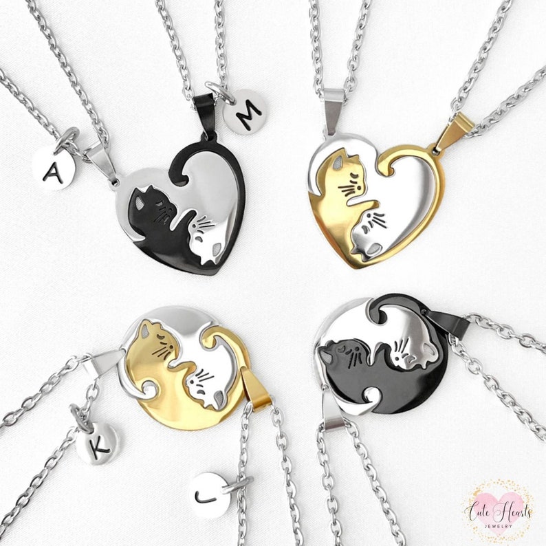 May include: Four sets of best friend necklaces with two matching pendants. Each pendant features a black and white cat and a gold and white cat. The pendants are shaped like a heart and a circle. The necklaces are silver chains with small round charms that have the letters A, M, K, and J engraved on them. The necklaces are displayed on a white background.