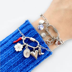 May include: Two silver charm bracelets with various charms, including a red chili pepper, a white flower, a heart, a boot, a sun, a pretzel, and a lock. The bracelets are on a blue knitted surface.