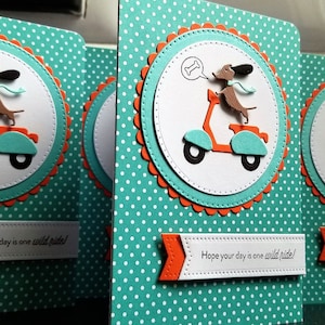 Könnte beinhalten: Grußkarten mit türkisfarbenem Polka-Dot-Hintergrund. Jede Karte zeigt einen weißen Kreis mit einem Dackel auf einem orangefarbenen Roller. Der Kreis wird von einem türkisfarbenen und orangefarbenen, gewellten Rand umrahmt. Der Text auf der Karte lautet "Hope your day is one wild ride!"