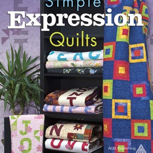 May include: A book titled "Simple Expression Quilts" by Fran Morgan. The book cover features a bookshelf with colorful quilts stacked on the shelves. The quilts have various patterns and designs, including letters, squares, and flowers. The book is published by AQS Publishing.