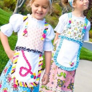 May include: Two young girls wearing colorful, patterned dresses with white aprons and headbands. The dresses have ruffles and pom-poms. The text "the Piccadilly jumper No.23" is visible on the bottom of the image.
