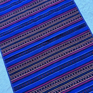 Può includere: Tessuto rettangolare blu con motivo a righe. Il design presenta bande alternate di blu, rosso, giallo e bianco, creando un aspetto vibrante e strutturato. Il tessuto è probabilmente un oggetto decorativo, come un runner da tavolo o un arazzo.