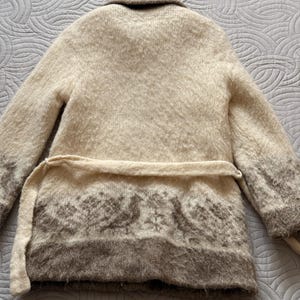 May include: A beige, long-sleeved cardigan with a shawl collar and a matching belt. The bottom of the cardigan features a brown patterned design. The cardigan is made of a soft, textured material.