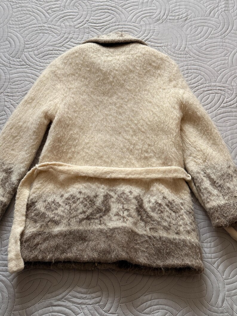May include: A beige, long-sleeved cardigan with a shawl collar and a matching belt. The bottom of the cardigan features a brown patterned design. The cardigan is made of a soft, textured material.