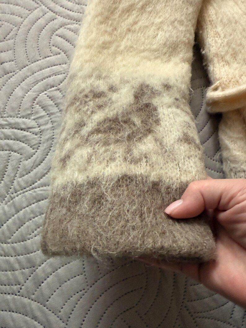 May include: Close-up of a beige and brown knitted sleeve. The sleeve has a textured, fuzzy appearance with a patterned design in the middle. The cuff is a darker shade of brown.