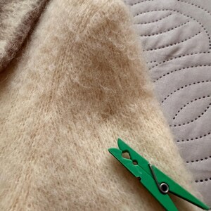 May include: Close-up of a cream-colored, fuzzy knit sweater. A bright green clothespin rests on the sweater's fabric. The sweater appears soft and textured, with a high neckline visible in the upper left corner.