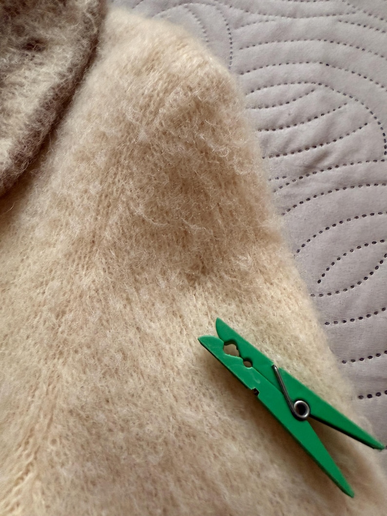 May include: Close-up of a cream-colored, fuzzy knit sweater. A bright green clothespin rests on the sweater's fabric. The sweater appears soft and textured, with a high neckline visible in the upper left corner.