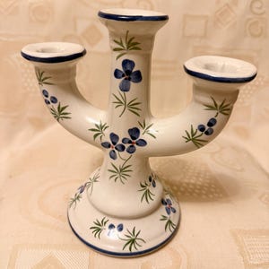 Peut inclure: Un bougeoir en céramique blanche à trois bras. Il est orné de motifs floraux bleus et de feuilles vertes. Les bords sont bordés de bleu. Un objet décoratif pour une table ou une étagère.