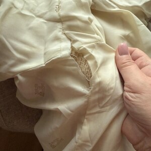 May include: Close-up of a cream-colored fabric with a tear revealing a woven texture underneath. The fabric has a sheen and appears to be part of a garment. The words "Deluxe Allround" are printed on the fabric.