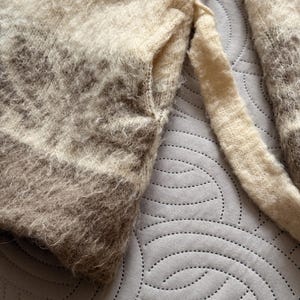 May include: Close-up of a soft, textured sweater in shades of cream, beige, and brown. The sweater has a fuzzy appearance with a pocket detail and a long sleeve. The color gradient creates a subtle striped effect.