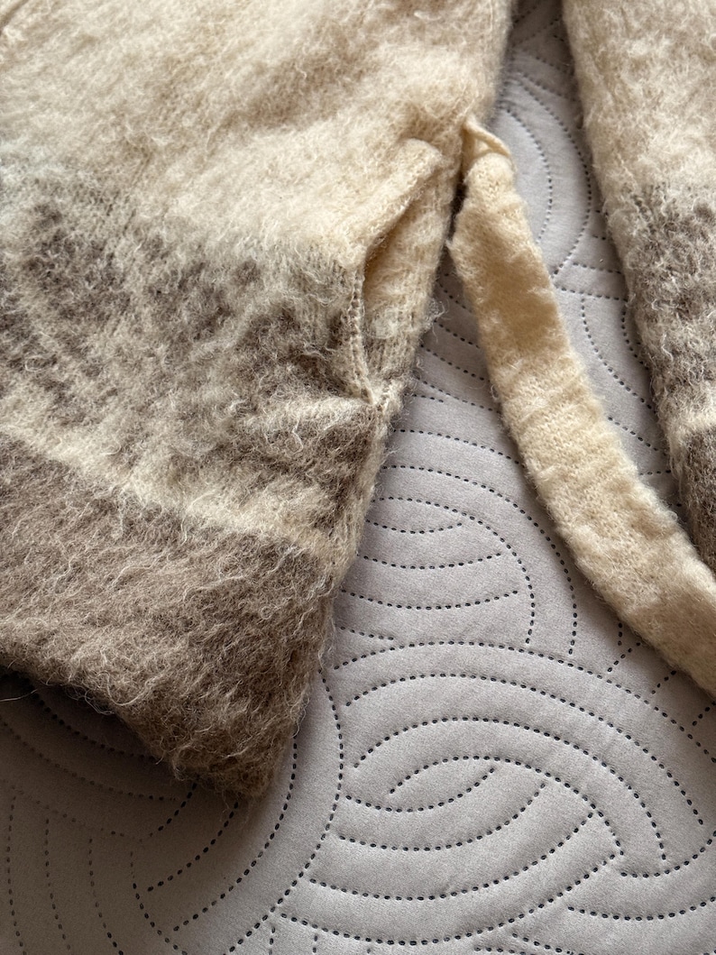 May include: Close-up of a soft, textured sweater in shades of cream, beige, and brown. The sweater has a fuzzy appearance with a pocket detail and a long sleeve. The color gradient creates a subtle striped effect.