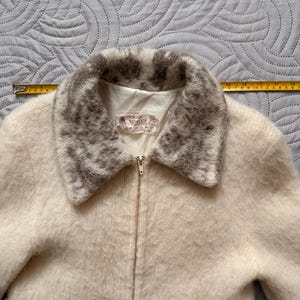 May include: Cream-colored zip-up jacket with a textured, fuzzy appearance. The collar is trimmed with a patterned gray and white design. The jacket has a label that reads "maloss keywool".