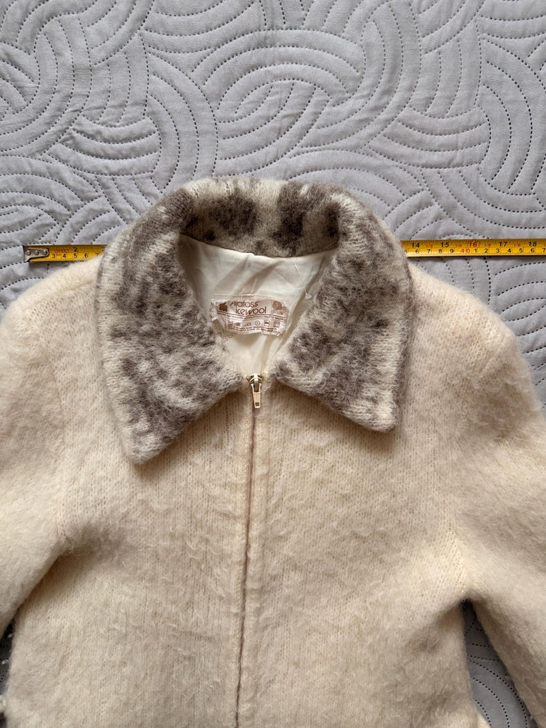 May include: Cream-colored zip-up jacket with a textured, fuzzy appearance. The collar is trimmed with a patterned gray and white design. The jacket has a label that reads "maloss keywool".