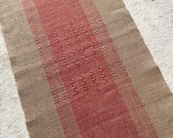 May include: A woven runner rug with a central red stripe and tan borders. The red stripe features a repeating pattern of small, raised rectangles. The rug has a textured appearance and is likely made of natural fibers.
