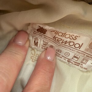 May include: Close-up of a label with the text "Alatoss icewool" and washing instructions. The label is brown and white, with the size "M" visible. The label is held by a person's hand.