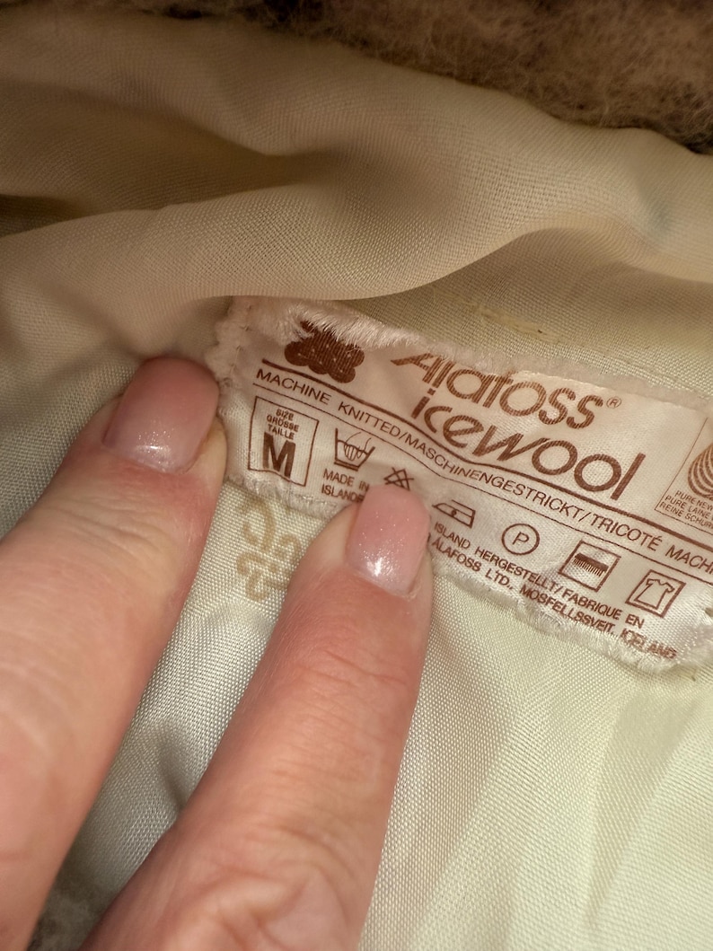 May include: Close-up of a label with the text "Alatoss icewool" and washing instructions. The label is brown and white, with the size "M" visible. The label is held by a person's hand.