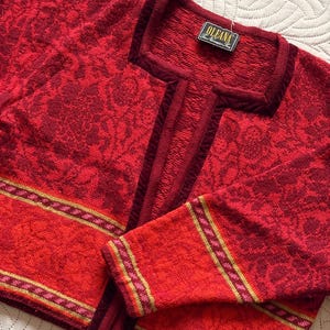 Peut inclure: Un pull rouge tricoté avec un motif floral et une encolure carrée. Le pull a une bordure rouge foncé contrastante et une bande orange avec des accents dorés et roses. L'étiquette indique "OLEANA The Norwegian Co."