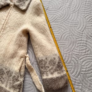 May include: A cream-colored, zip-up coat with a fuzzy texture and a gray collar and cuffs. The coat has a matching belt and a label that reads "House of Wool". A yellow measuring tape is next to the coat.