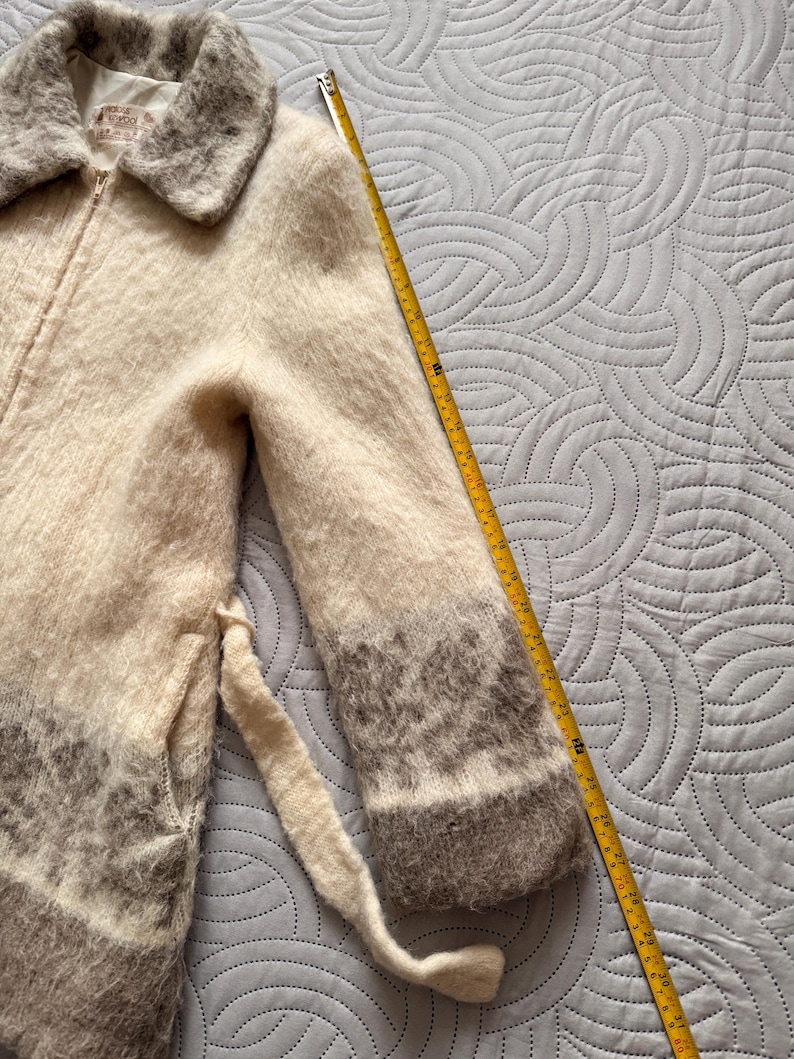 May include: A cream-colored, zip-up coat with a fuzzy texture and a gray collar and cuffs. The coat has a matching belt and a label that reads "House of Wool". A yellow measuring tape is next to the coat.