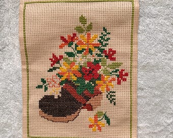 May include: A cross-stitch embroidery depicting a brown boot filled with vibrant flowers. The flowers are in red, yellow, orange, and green, set against a beige background with a green border. The artwork is a handmade piece.