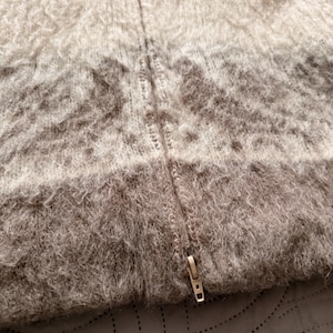 May include: Close-up of a textured, gradient-colored sweater with a zipper. The sweater transitions from a light beige at the top to a darker brown at the bottom. The zipper is a metallic gold color.