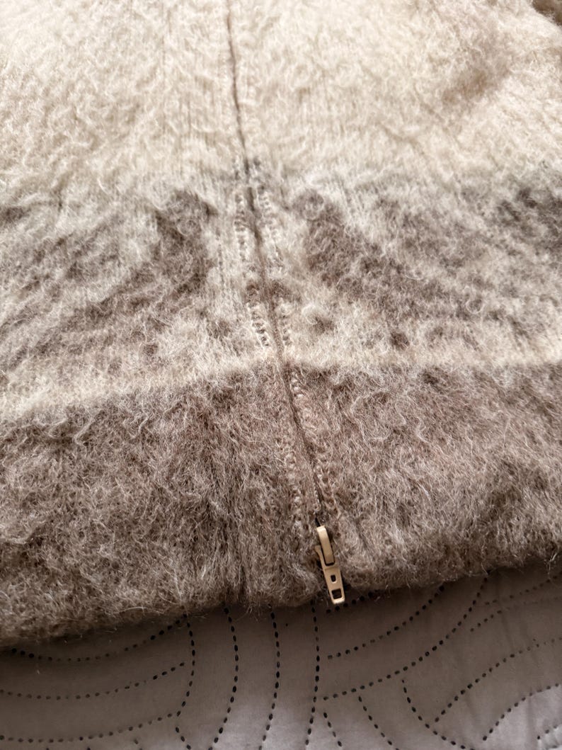 May include: Close-up of a textured, gradient-colored sweater with a zipper. The sweater transitions from a light beige at the top to a darker brown at the bottom. The zipper is a metallic gold color.