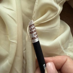 May include: Close-up of a cream-colored fabric with a tag attached. The tag reads "FUTT LIN DOUBLE 100 POLY". A black object is held in the hand, partially obscuring the fabric.