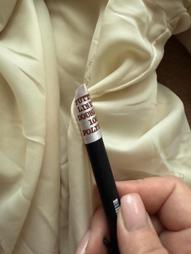 May include: Close-up of a cream-colored fabric with a tag attached. The tag reads "FUTT LIN DOUBLE 100 POLY". A black object is held in the hand, partially obscuring the fabric.