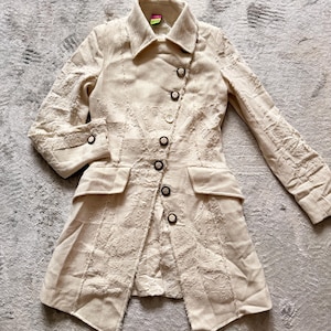 May include: Beige linen jacket with a unique asymmetrical button closure. The jacket features a notched collar, two front pockets, and decorative stitching. The long sleeves have buttoned cuffs. The jacket is approximately 30 inches long.