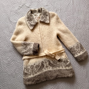 May include: A cream-colored coat with a zip-up front and a matching belt. The coat features a patterned design in shades of brown on the cuffs, collar, and lower hem. The coat is made of a soft, textured material.