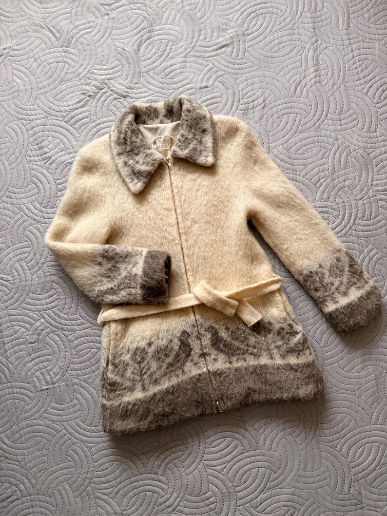 May include: A cream-colored coat with a zip-up front and a matching belt. The coat features a patterned design in shades of brown on the cuffs, collar, and lower hem. The coat is made of a soft, textured material.