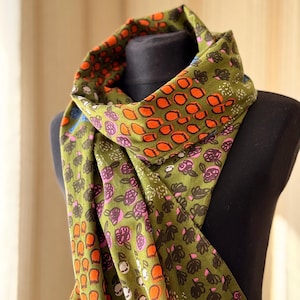 May include: Olive green scarf with a repeating pattern of orange and pink floral and abstract shapes. The scarf is draped over a black mannequin, showcasing the fabric's design and drape. The scarf's design includes orange, pink, and white accents.