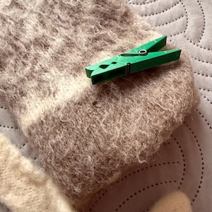 May include: Close-up of a light brown and cream-colored knitted scarf with a green clothespin. The scarf has a textured, fuzzy appearance. The clothespin is made of green plastic.