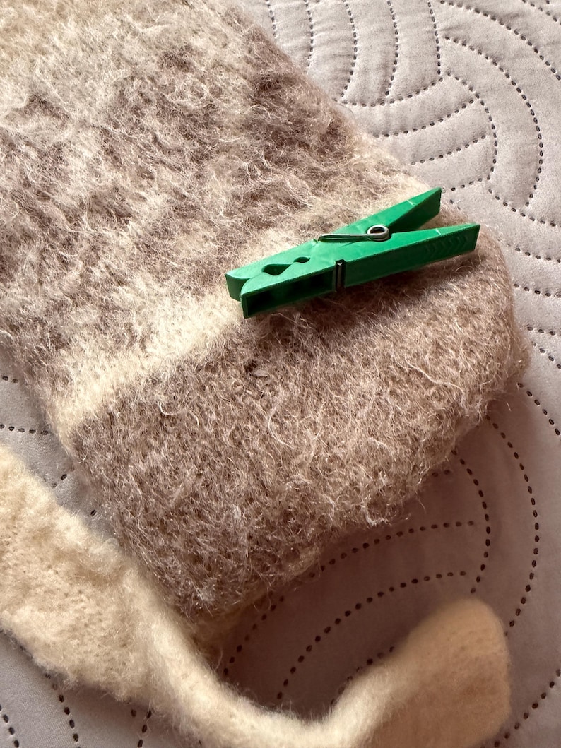 May include: Close-up of a light brown and cream-colored knitted scarf with a green clothespin. The scarf has a textured, fuzzy appearance. The clothespin is made of green plastic.