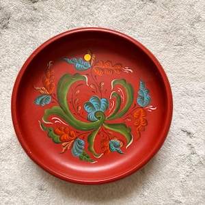 Pode incluir: Uma bandeja redonda de madeira vermelha com um design floral pintado à mão. O design apresenta folhas e flores verdes, azuis e laranja. A bandeja é uma peça decorativa, provavelmente para servir ou exibir.